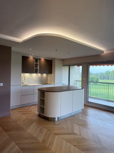 Cuisine moderne avec îlot central, éclairage doux et grande fenêtre donnant sur un paysage verdoyant.